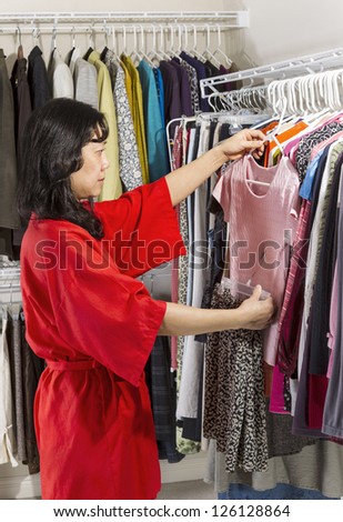 Vertical portrait of mature Asian woman, dressed in red bath robe, in walk-in closet coordinating her clothes