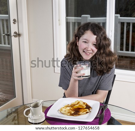 Young adolescent girl drinking milk with big smile on her face and single large pancake on white plate with maple syrup on top