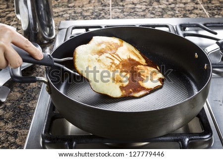 Horizontal photo of main focus on pancake being flipped in hot frying pan on top of gas stove range