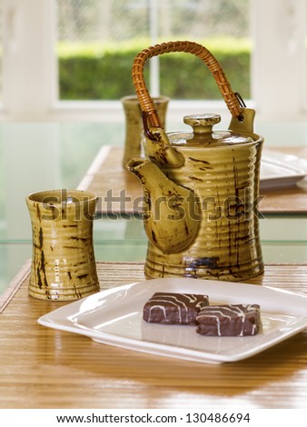 Vertical photo of tea pot, cup, white plate with cookies on bamboo place mat in family formal dining room with daylight coming through window in background
