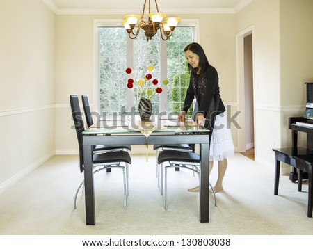 Photo of mature woman placing diner mats in family formal dining room table with daylight coming through large windows in background