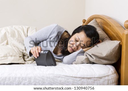 Horizontal photo of mature woman, lying head down in pillow, while holding alarm clock in other hand