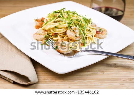 Closeup horizontal photo of Pasta dish with focus on large shrimp in stainless steel fork, basil, cloth napkin, red wine, and parsley in dish