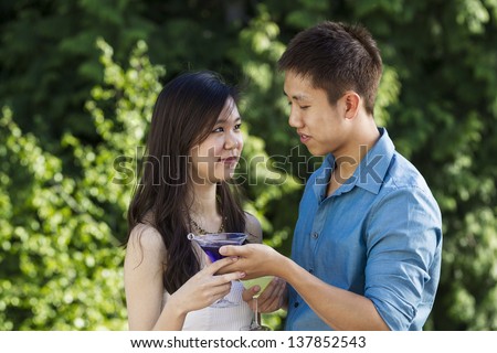 Horizontal photo of a young adult man handing his lady friend a drink while outdoors with green trees in background