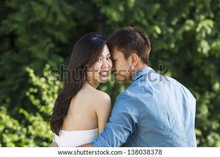 Horizontal photo of a young adult couple holding each other with lady friend looking behind her with green trees in background
