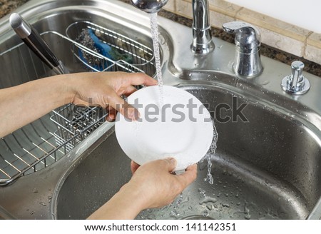 Horizontal photo of female hands rinsing off a small white dinner plate with kitchen sink and running faucet in background