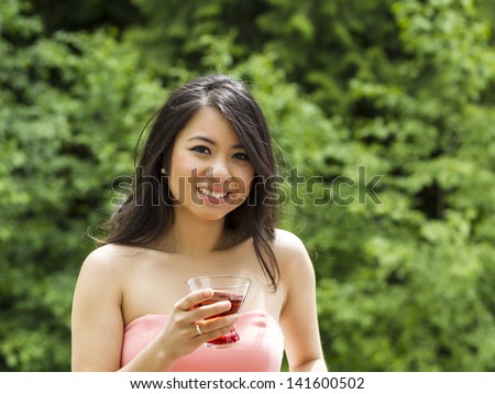 Horizontal photo of young Adult woman looking forward while holding drink outside with blurred green trees in background