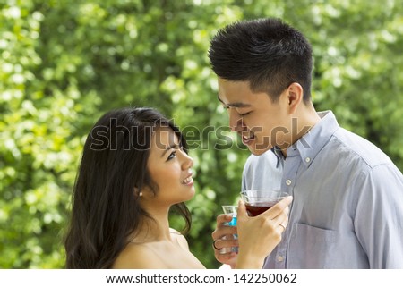 Horizontal photo of young adult couple smiling at each other while holding mixed drinks with blurred out green trees during daylight in background