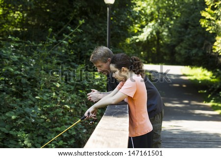 Horizontal photo of young girl and her father fishing off wooden bridge with trees and lamp post in background