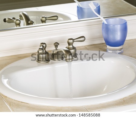 Photo of white bathroom sink and running faucet with blue cup, tooth brush and mirror in background