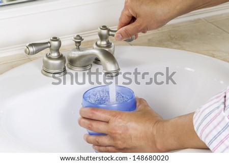 Horizontal photo of female hands putting water into blue cup with white bathroom sink and faucet in background