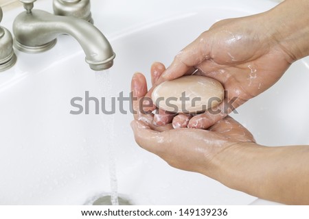 Horizontal photo of female hands lathering up with bar of soap, running water and bathroom sink in background