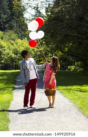Vertical photo of young adult couple dressed in formal attire looking at several balloons while holding picnic basket with walking path, green grass and trees behind them