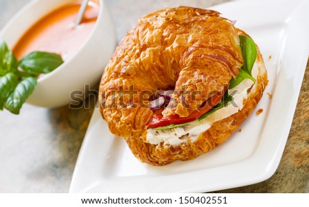 Horizontal photo, slightly angled, of chicken salad, tomato, and onion inside croissant bread on white square plate with tomato soup, basil and spoon in bowl all resting on natural stone counter top