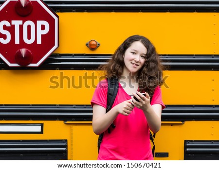 Horizontal photo of young girl standing by side of bus, with back pack over her shoulders, cell phone in hands while looking forward
