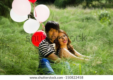 Horizontal photo of young adult couple, sitting in the middle of a tall grass field, with several white and red balloons while expressing love by holding each other