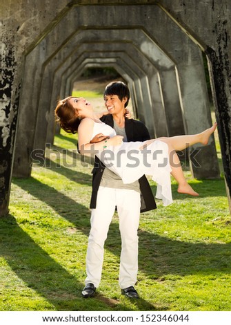 Vertical photo of young adult man holding his lady in his arms while the laugh together underneath a structured column wall in background