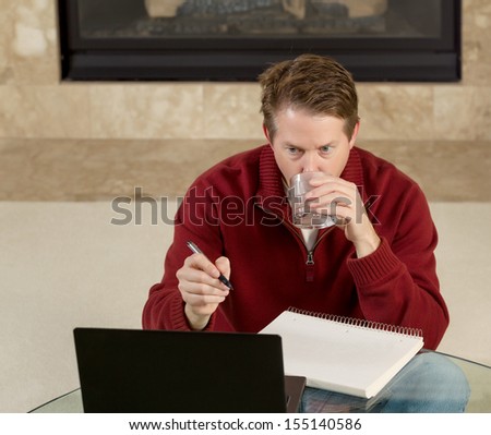 Photo of mature man, drinking water while sitting down at glass table, working from home, looking at computer screen with fireplace in background