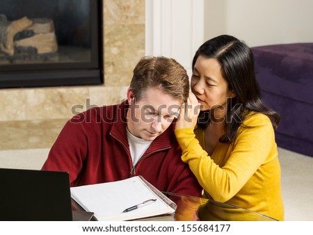 Photo of mature couple, with woman whispering into ear of man, while working from home with fireplace and partial sofa in background