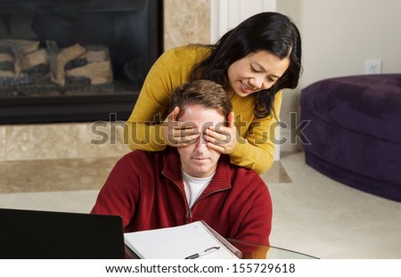 Photo of mature couple, with woman covering up the eyes of man, while working from home with fireplace and partial sofa in background