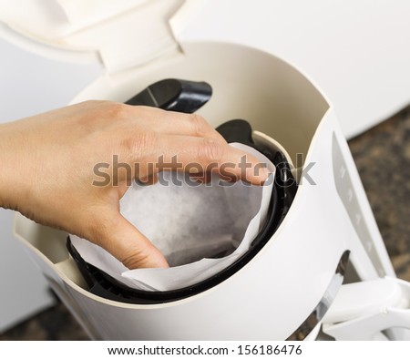 Angled photo of female hand putting in white bleached coffee filter with coffee maker, and stone kitchen counter top in background