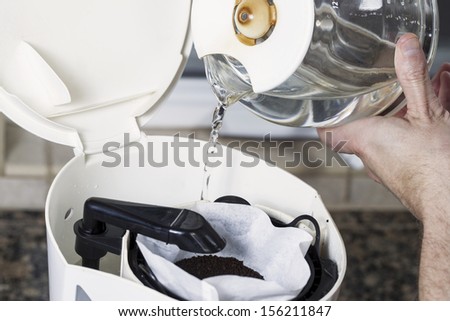 Horizontal photo of male hand pouring water, from coffee pot, into coffee maker with fresh coffee grounds in white bleached coffee filter with stone kitchen counter top
