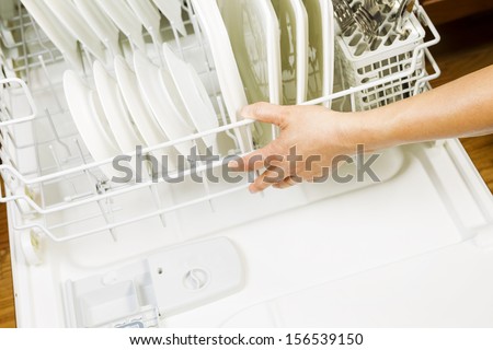 Horizontal photo of female hand closing lower dishwasher rack that is filled with white plates, stainless steel spoons and forks with red oak floors in background