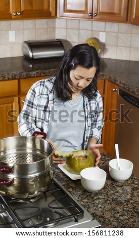 Vertical photo of mature woman putting top of cooked winter melon back on while placed in dinner plate next to cooking kettle on stove top