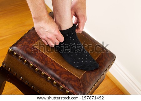 Horizontal photo of man pulling up dress sock while placing foot on padded foot stool with red oak floors and white wall in background