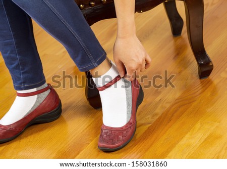 Horizontal photo of woman snapping strap on causal shoes while sitting on leather padded footstool with red oak floors in background