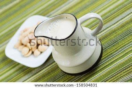 Angled horizontal photo of almond milk in pitcher with fresh almonds, in a white dish, with green table cloth in background