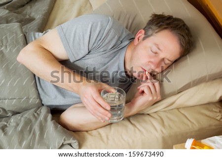 Horizontal photo of mature man putting medicine pill to his mouth while holding glass of water in bed