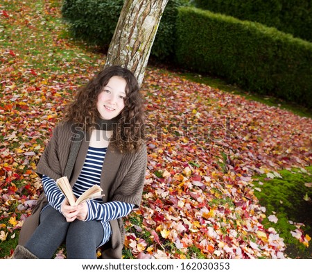 Photo of teenage girl, looking forward, enjoying her book while lying against a tree during the autumn season dressed in earth colors