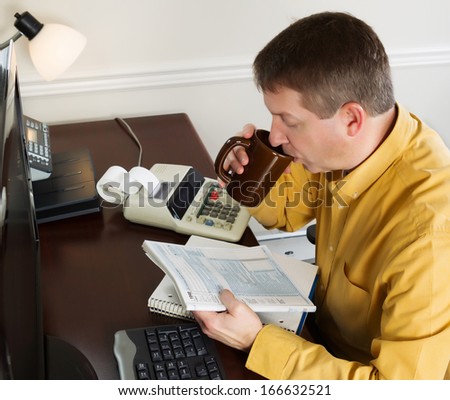 Horizontal photo of mature man looking over the tax tables while sipping on coffee in his office at home