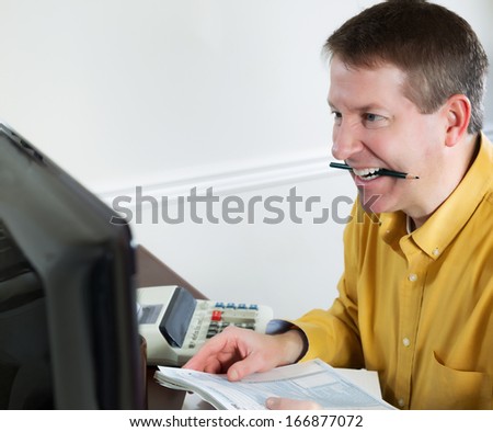 Photo of mature man, looking at data on computer monitor, while biting on his pencil in anger, with office equipment in background