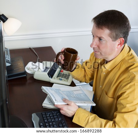 Photo of mature man, looking at data on computer monitor, working on his taxes while holding cup of coffee with office equipment in background