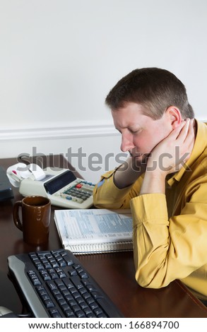 Vertical photo of mature man, holding head with hands, working on his taxes with tax booklet and office equipment in background