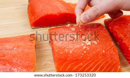 Horizontal photo of female fingers placing sea salt on fresh pieces of Wild Red Salmon and natural bamboo board in background