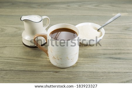 Closeup horizontal photo of a full cup of coffee with sugar and spoon in bowl and cream in pouring spout with aged wood underneath