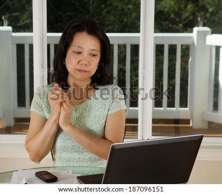 Photo of mature woman looking at computer screen while working at home with laptop, cell phone and papers on top of table and large windows in background