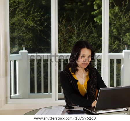 Photo of mature woman listening to music while working at home with laptop, cell phone, calculator and papers on top of table and large windows in background