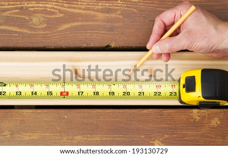 Horizontal photo of hand holding pencil next to tape measure on top of new cedar wood board next to fading wood on outdoor wooden deck