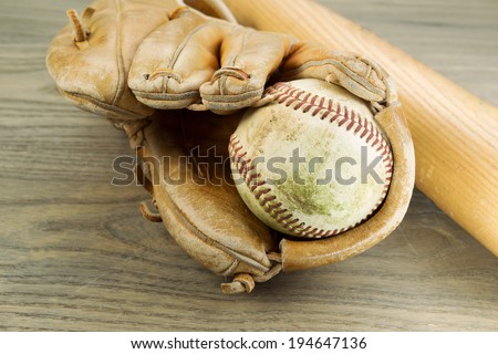 Closeup horizontal photo of an old dirty baseball inside of heavily used glove and wooden bat in background on rustic wood