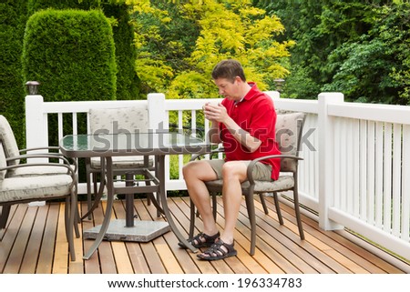 Horizontal photo of mature man holding coffee of hot coffee while preparing to read a magazine on outdoor patio with green and yellow trees in full seasonal bloom
