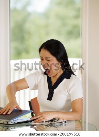 Vertical image of mature woman angrily slamming her laptop shut while working from home with bright daylight coming in from window in background