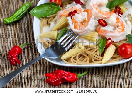 Closeup front view of stainless steel fork holding single shrimp with Alfredo Pasta dinner placed on white plate and natural bamboo mat underneath