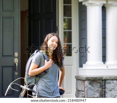 Image of teenage girl, waist up and looking forward, carrying school bag while resting against her bicycle with home in background