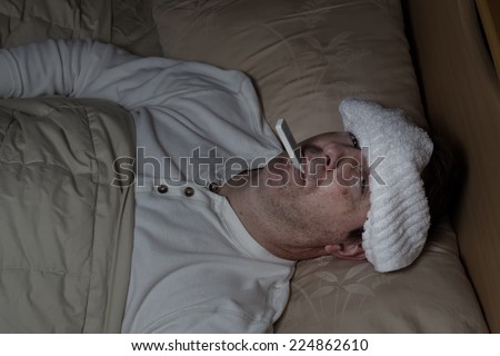 Horizontal image of mature man, with wet towel on forehead, lying down in bed testing his temperature