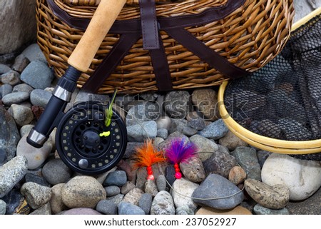 Top view up of  fishing fly reel, landing net, creel and assorted flies on dry river bed rocks
