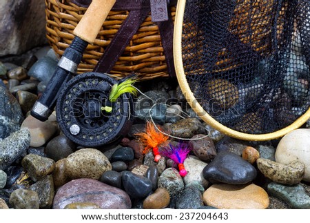 Closeup of fishing fly reel, landing net, creel and assorted flies on wet river bed stones